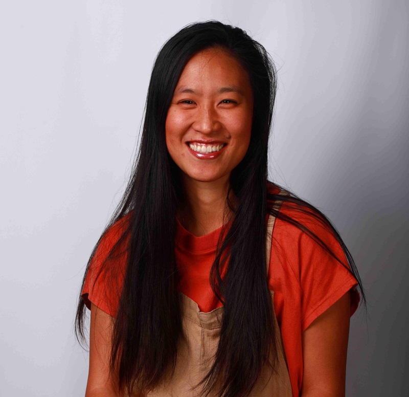 Yumi Inoue is a japanese descendent woman, with long and black hair. She is smiling and wearing an orange T-shirt and beige rompers.